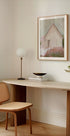 a modern, minimalist workspace with a wooden desk, a woven chair, a lamp, and a framed painting of a house.