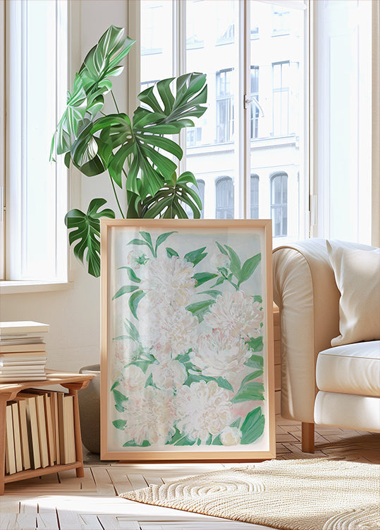 a large framed print of white flowers with green leaves, placed on a wooden floor in a bright, airy room with large windows.