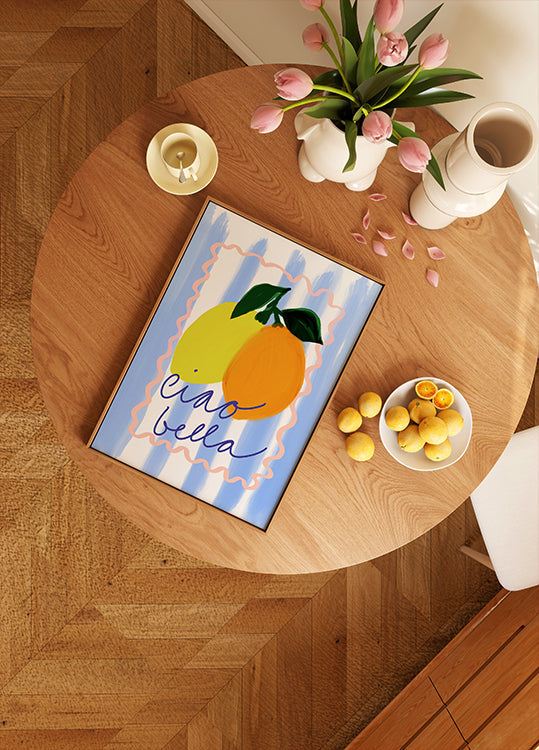 a wooden table with a framed artwork depicting two lemons, a bowl of fruit, and a vase of pink flowers.