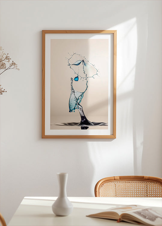 a framed artwork of a water droplet suspended in mid-air, with a blue and white color scheme. The artwork is displayed on a white wall above a table with a white vase and a book.